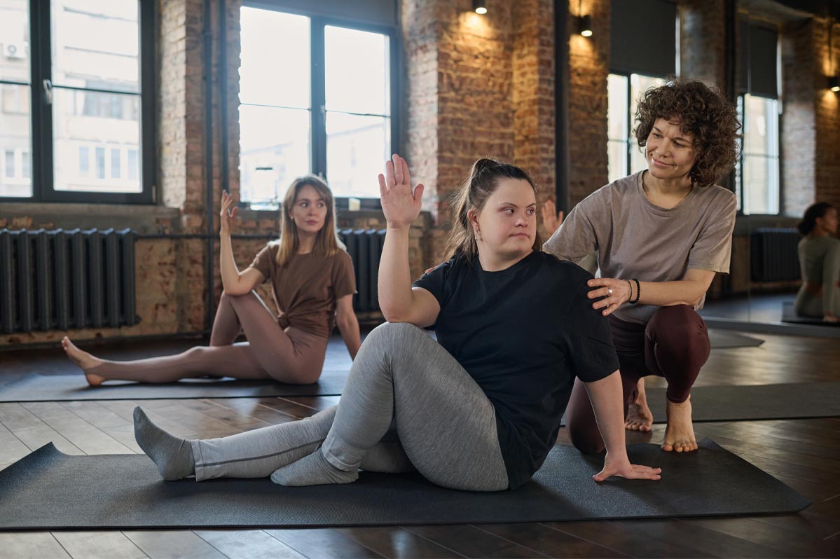 Three women are in a yoga studio. The woman (Beth) in the center is setting on yoga matt, sitting up, with her right leg straight and the left leg bent. Her right elbow is on her left knee, and her hand is pointing up while her body and head is turned to her left. She has down syndrome. Another woman is kneeing behind her, touching her left shoulder and her back to ensure the correct position. The other woman is the background is in the same pose as the women participating in the exercise. All three women are in yoga pants and tops.