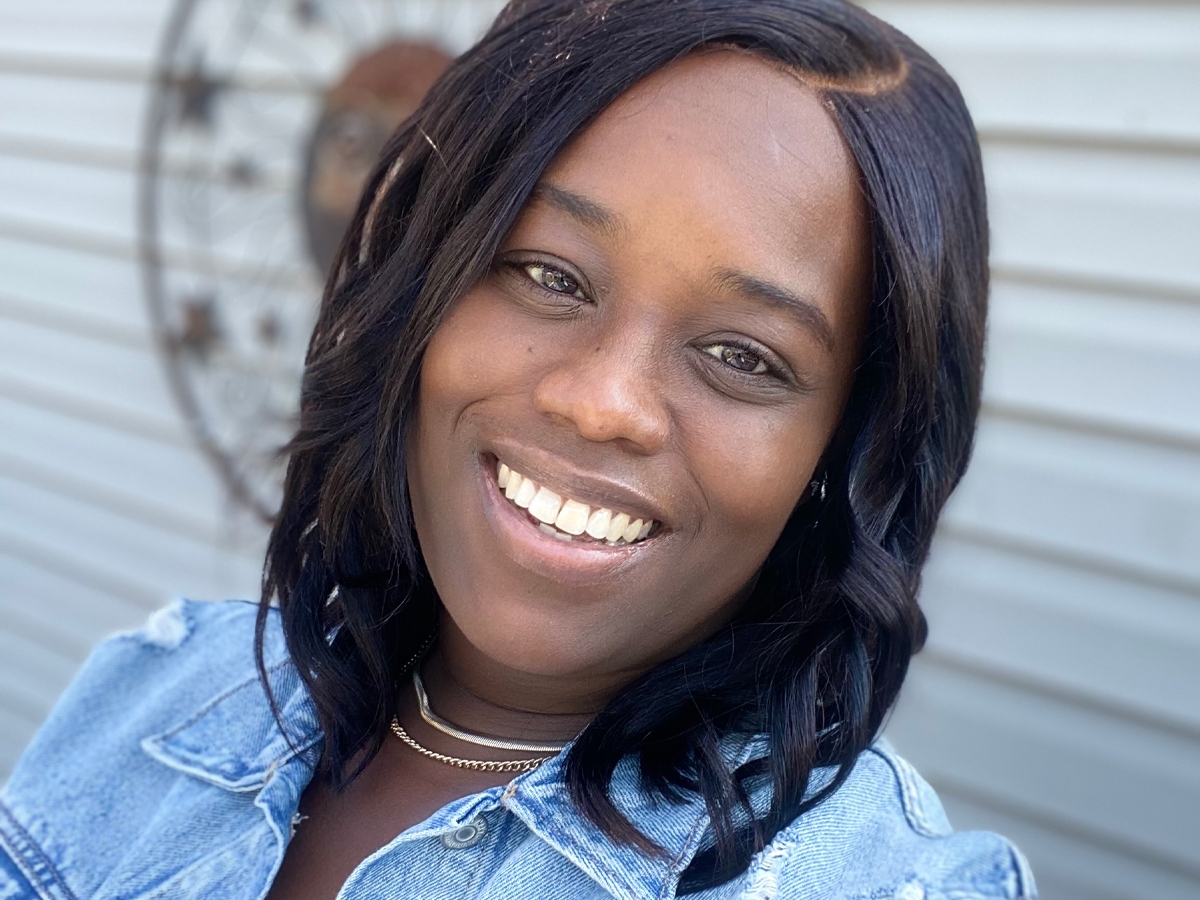 Person is outside looking straight at the camera smiling, looking right at the camera, with dark shoulder length hair, brown eyes, with a smile, blue jean jacket, and a dark top with white strips.