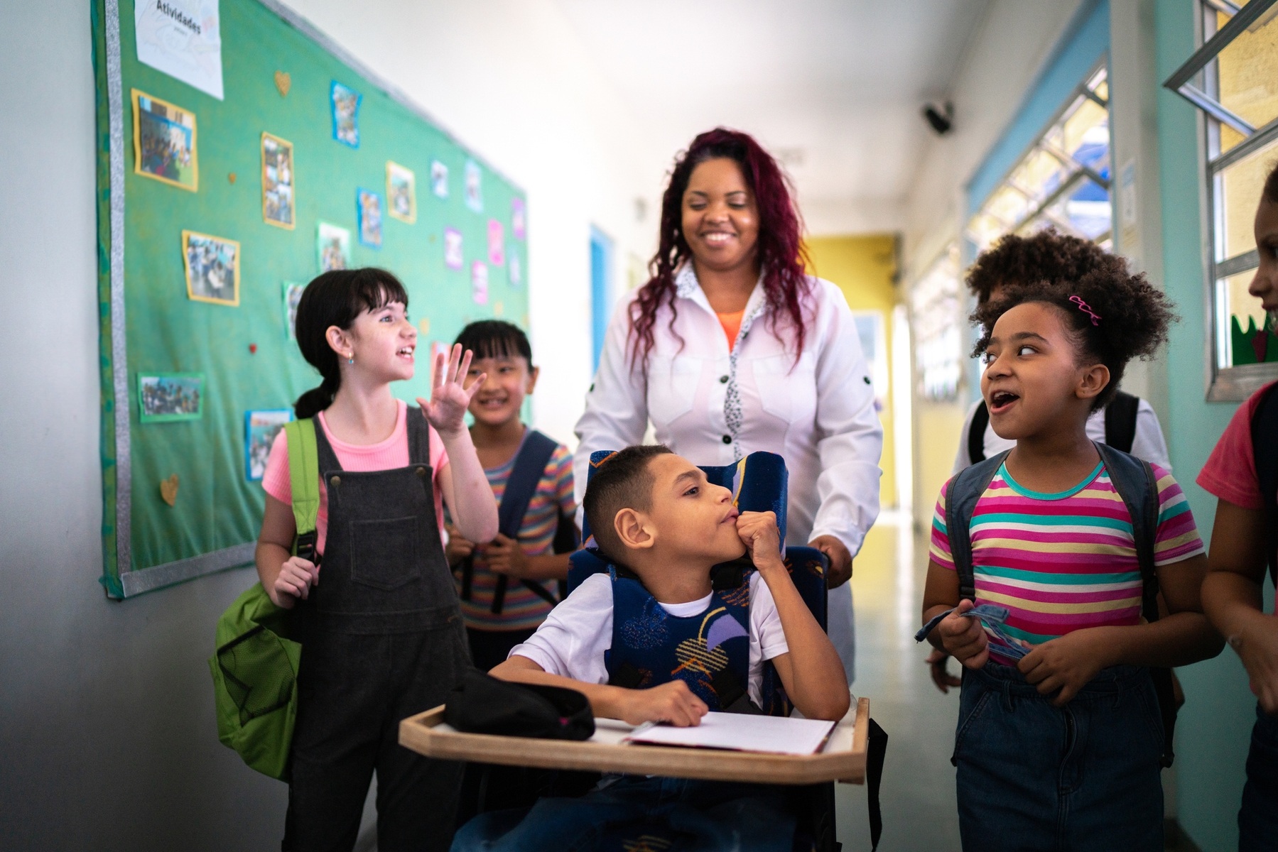 A female paraprofessional pushes a child's wheelchair down a school hallway. They are surrounded by a small group of his classmates, who talk excitedly.