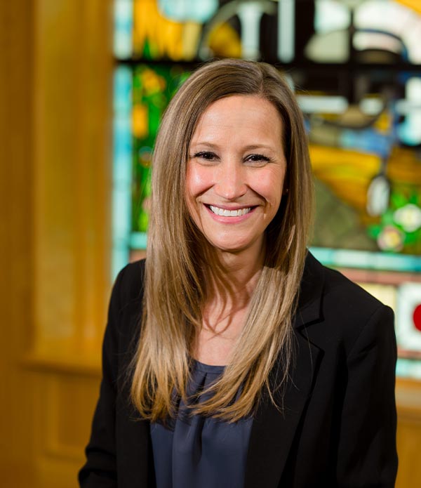 Brooke Graff is smiling at the camera. She has long light brown hair, is wearing a navy-blue blouse and a black jacket with the sleeves rolled up. She is standing in front of a stained-glass window.