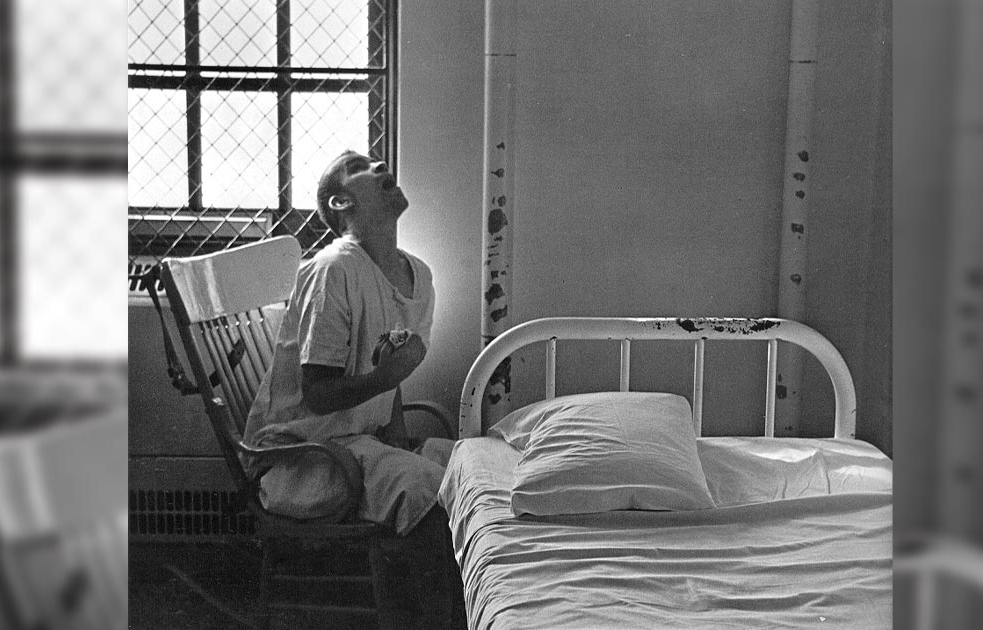 A black-and-white photo of a man in a hospital gown, clasping his chest and staring at the ceiling. He sits in a rocking chair. A harness is tied to the back of the chair. A steel grid covers the closed window behind him. His knees touch a narrow, institutional bed with a steel-framed headboard. Two steel bars are between the headboard and the wall. There are wear marks and nicks on the steel bars and headboard, which are painted white. The bed sheets are also white. The rest of the room is bare.